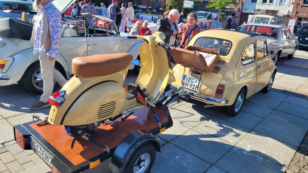 Ein Oldtimer mit offenem Kofferraum und ein Oldtimer-Roller auf einem Anhänger, geparkt zwischen anderen Oldtimern. Mehrere Personen haben sich in einer sonnigen Umgebung im Freien um die Fahrzeuge versammelt.